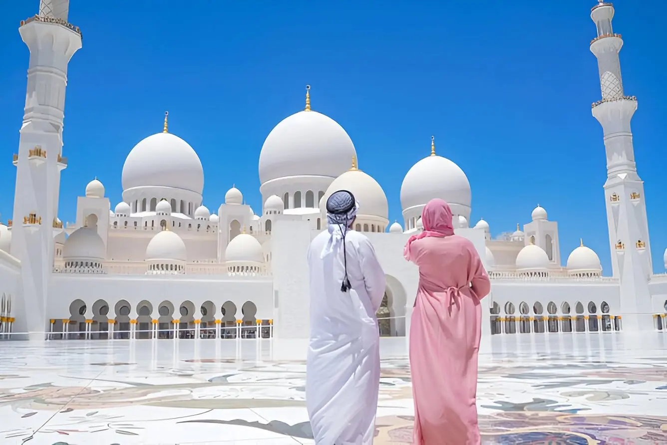 Two visitors in traditional dress looking at the white domes and minarets of Sheikh Zayed Grand Mosque in Abu Dhabi under a clear blue sky