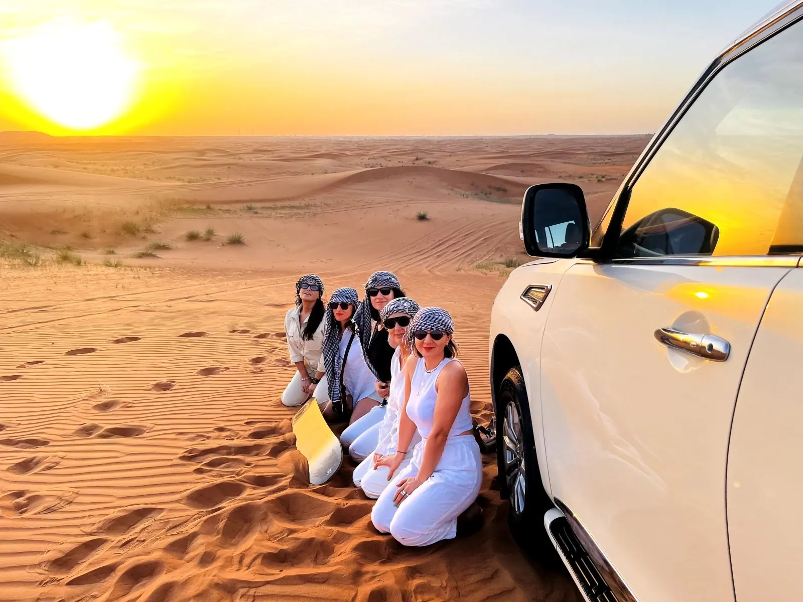 Group of travellers in checked headscarves kneeling on red sand dunes beside a white 4x4 at sunset on a Dubai desert safari