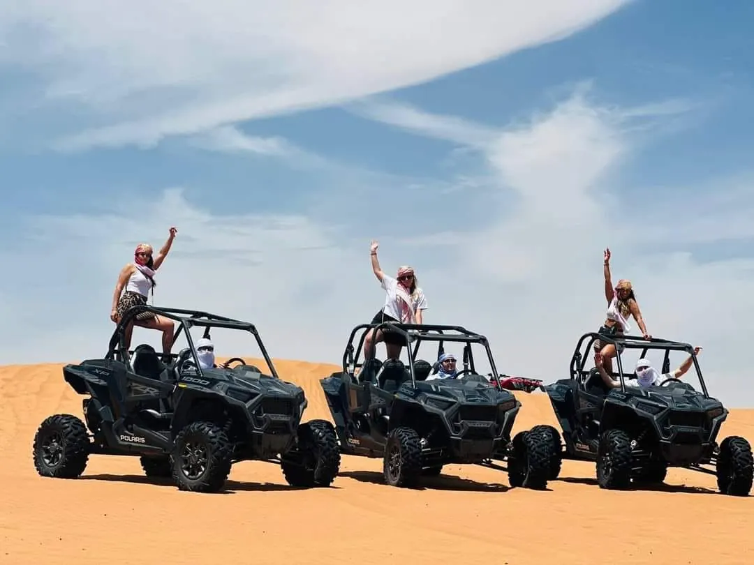 Group of friends waving from three black dune buggies lined up on a sand dune under a blue sky in the Dubai desert