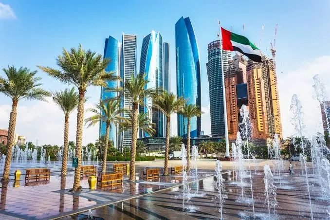 Etihad Towers and the Abu Dhabi skyline behind palm trees, water fountains and a waving UAE flag on a sunny day